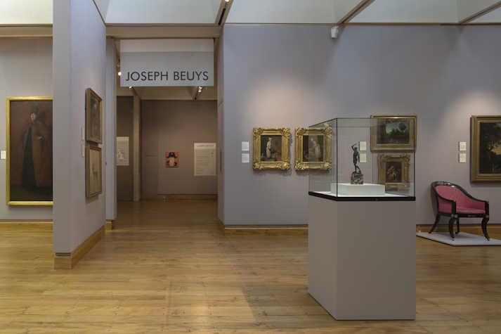installation view of a gallery. on the wall it reads 'Joseph beuys'. surrounding there are gilded framed works on a wall and a glass display case/plinth