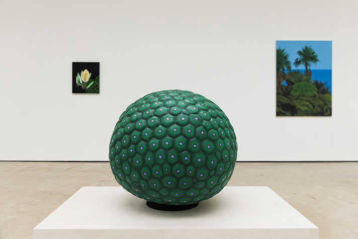 A large round, green sculpture sits on top of a white plinth in a white cube gallery. 2 Paintings are hung behind, on either side of the sculpture.
