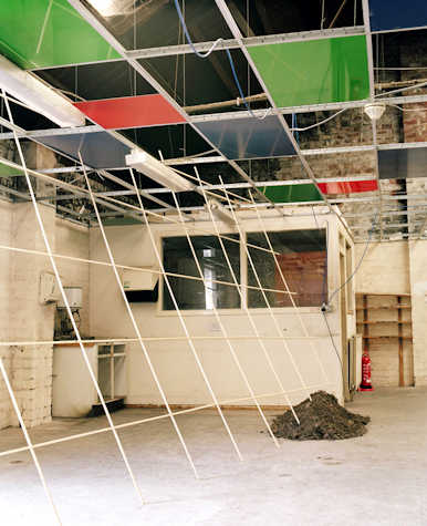 grid structure hanging from a ceiling with alternating red, green and blue tiles. leaning from this grid structure at an angle is another made of poles that reaches the floor. on the floor is a pile of dirt and rocks.
