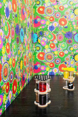 The image shows a colourful exhibition with bright patterned wallpaper and sculptures made of glass jars