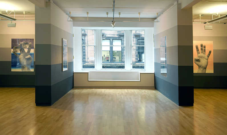 Art gallery with wooden floors and horizontally stripes in the walls in a gradiant from black to white from bottom to top. Windows in the centre, columns to the sides. Two similar artworks to each side of hands.