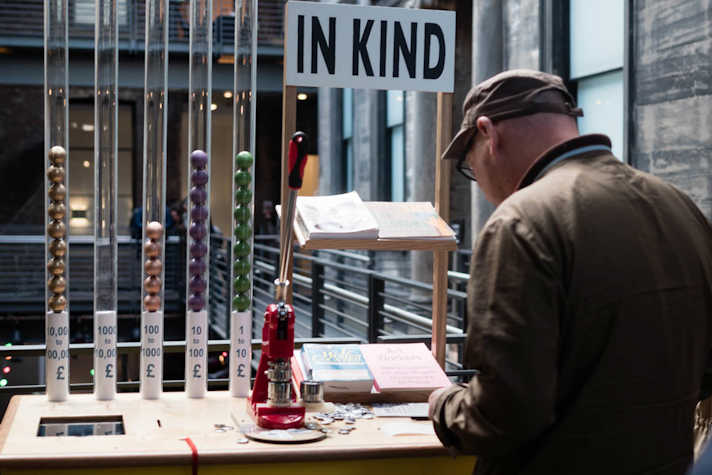 In the CCA a white man with a brown hat and jacket and glasses stands in front of a table that has a poster that reads "IN KIND" and has several pamphlets and books. There is also a pin-making machine and several tubes with colored balls in them and tags with quantities of money in them.