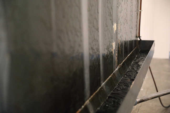 A close up of a dark sheet of metal, at the bottom is a trough that is gathering a liquid that runs down the sheet of metal.