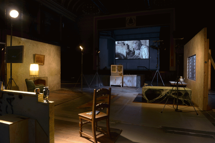 There are several movie sets facing each other, the closest one has a wooden chair. There is a screen in the back wall showing a black-and-white film. It is all illuminated by big spotlights with a warm yellow light. This is part of an art installation in Film City, Glasgow International 2018.