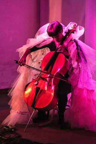 A person plays a cello wearing a large net costume. The light around the person is pink