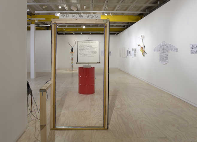 Art gallery installation featuring a red barrel, a framed sign labeled "ARTS MEDIA SPORTS," and various wall-mounted mixed-media pieces, including text-covered fabric and sculptural elements.