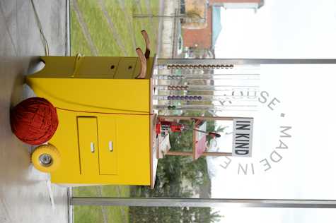 In Easterhouse a yellow trolley/information booth with a big ball of red yarn coming out of it with a poster that reads "IN KIND" and several pamphlets and books. There is also a pin-making machine and several tubes with colored balls in them and tags with quantities of money in them. It is in front of a glass panel that looks to a green garden.