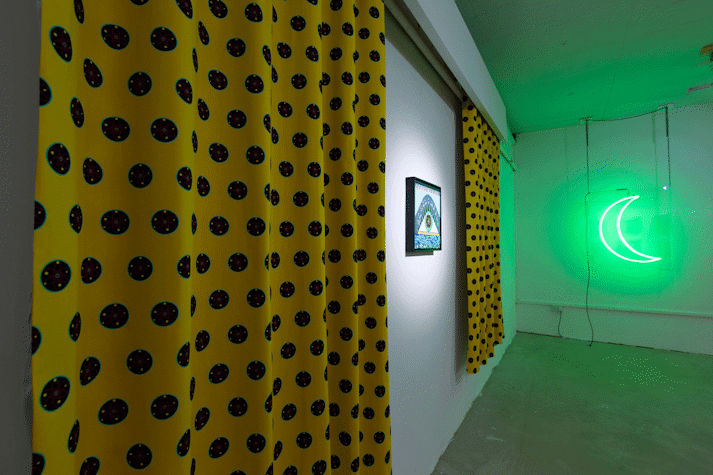 a small painting is hung on the wall and spot-lit, on it are abstract shapes and the words 'my heart is broken', a large curtain hangs either side with a repeat pattern of polka dots, in the background is a green neon light in the shape of a crescent moon