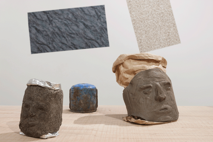 On a wooden surface, there are three clay sculptures. The two at the front are shaped like faces. Behind them the wall, has two textured rectangles printed on it.