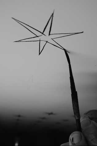 A black and white photograph showing the outline of a six-pointed star being painted in black on a white ceiling. The artist’s hand is visible in the foreground of the image and the bristles of his brush are gently compressed against the ceiling.