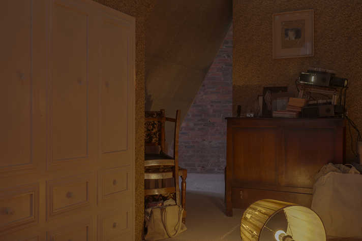 The corner of a dimly lit room. There are two chairs upturned and stack on each other, a wooden cabinet and a wardrobe, along with books and photographs.