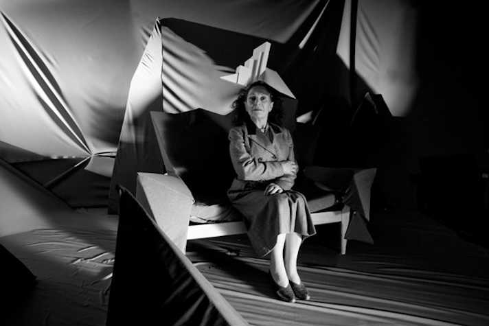 black and white film still of a woman sat on a bench with fabric hanging sculpturally behind