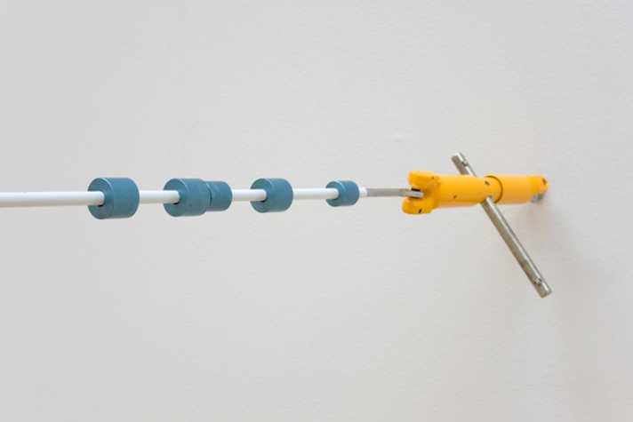 sculptural installation affixed horizontally to wall, rubber-coated wire cable with blue rings around it and a yellow clamp at the end closest to the wall.