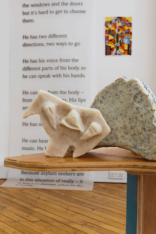 Sculptures are displayed on a wooden table. In the background there is artwork displayed on the wall featuring written text.