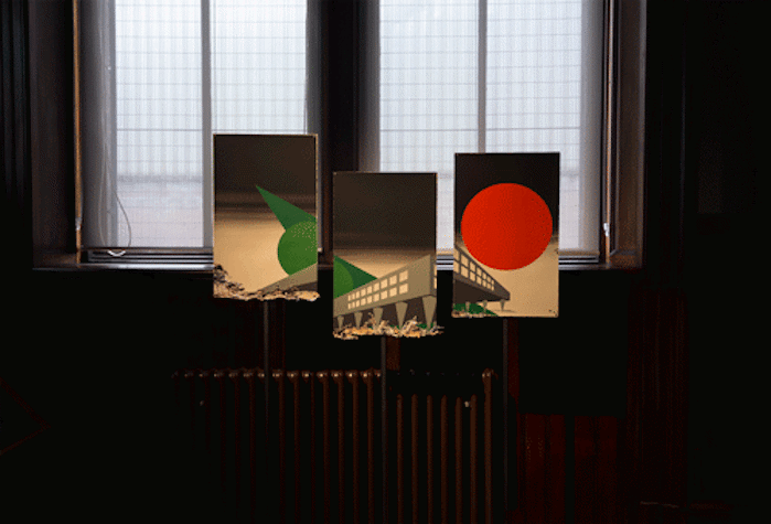 In a dark room, with windows with light behind and a radiator below, we see three rectangular painitngs that make up one in different altitudes. There is a grey building with a lot of windows and a green pointy shape to the left and a red circle to the right.