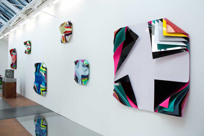 Installation view of colorful, abstract wall sculptures with layered, folded surfaces on a white gallery wall.