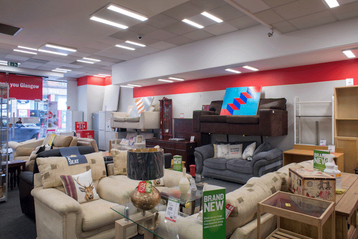 In a British Heart Foundation charity shop with earth toned furniture there are two paintings of colorful stairs.