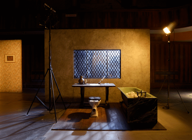 A wooden board floor has on top a wooden desk and a toilet as a chair with a green bathtub to the right. There is a wall with a window with diamond-shaped metal bars. It is all illuminated by two big spotlights with a warm yellow light. There are other sets behind and around this one. This is part of an art installation in Film City, Glasgow International 2018.