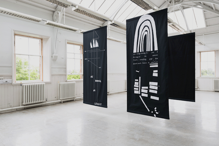 four portrait orientation large black rectangles of fabric hang from the rafters of a large school hall-esque room. The front two have white printed text, diagrams and shapes on.