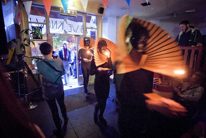 three performers in all black with large pleated collars holding torches enter a room from outside as onlookers watch.