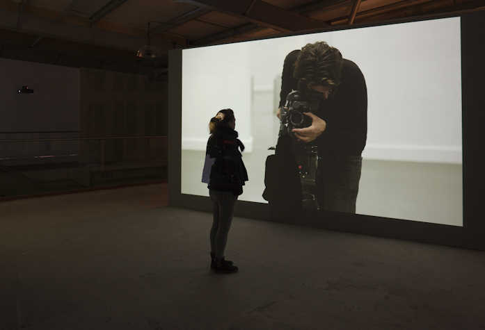 a person is stood in front of a large projection screen. on the screen there is a person looking into a viewfinder taking a photograph