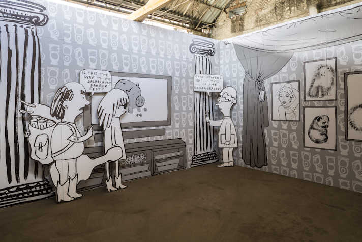 We can see two walls intersecting in a corner, both with drawn black and white murals. Left wall that has two Greek columns encasing a screen with a piggy bank. One person, with a backpack and a guitar, is asking "Is this the way to the Dalmarnock danger fest?". Besides him is a person with long hair and sunglasses. The third person, to their right, answers "God heavens no!.... this.... is the regenerators", while touching one of the Greek columns. On the right wall there is a curtain and paintings.