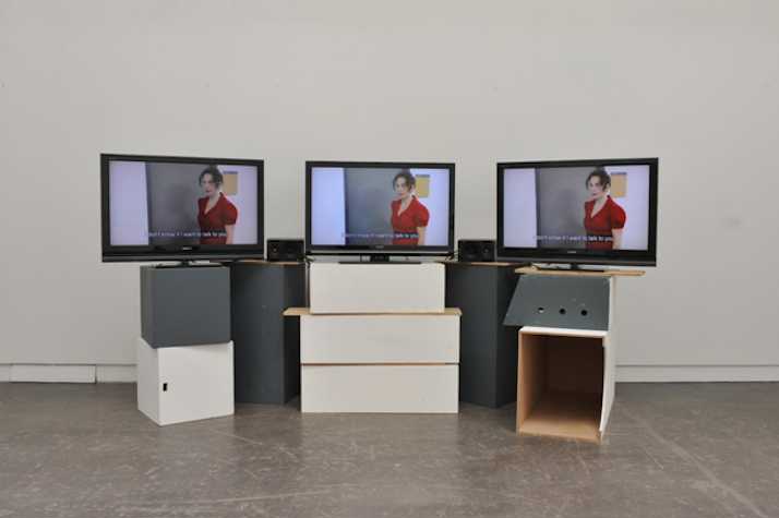 3 TV monitors playing the same video, resting atop a pile of white and grey wooden boxes