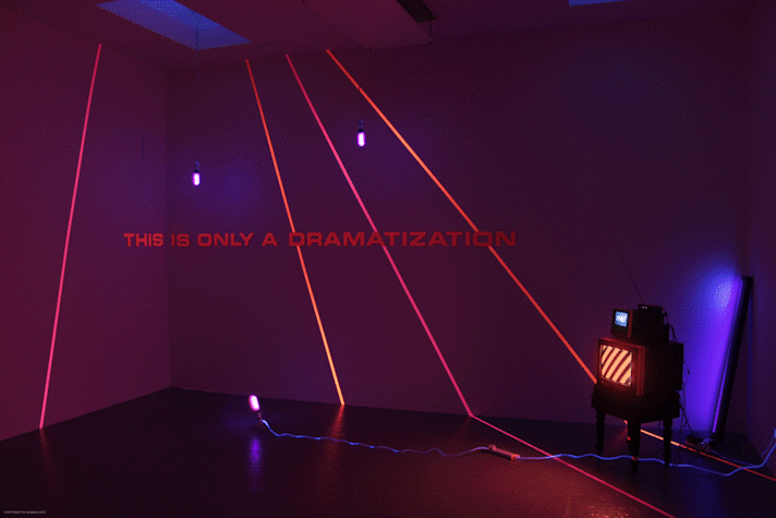 dark room with red and blue lighting. red laser like lines are projected across the wall along with he words 'THIS IS ONLY A DRAMATIZATION'. in the corner of the room is a small cube monitor with an even smaller monitor resting on top.