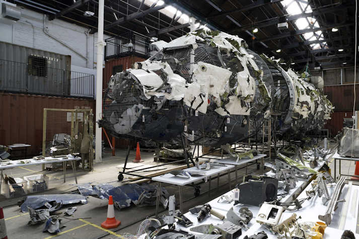 Interior of Tramway 2 with Wreckage of an aircraft reassembled as part of an art installation in an industrial space, with metal fragments, tables displaying parts, and shipping containers in the background.