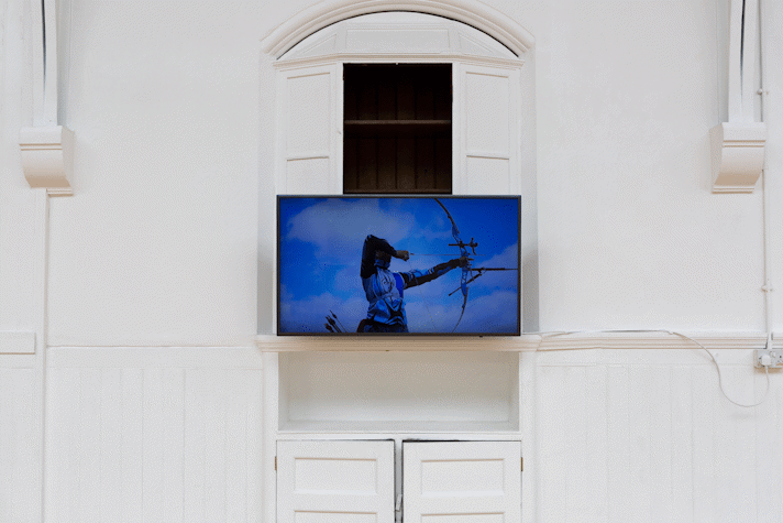 A TV is mounted on a white wall with wood panelling and period features. The image on the screen shows a person with a bow and arrow, pulling the arrow back.