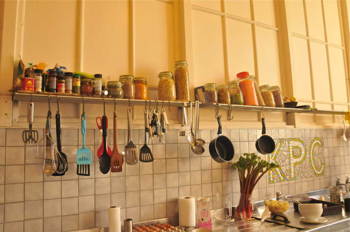 In the corer of a kitchen, there is a rack of containers and condiments. There are utensils and cutlery hanging from the rack. On the counter below that, there are many kichen supplies.