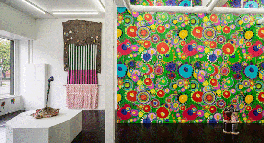 The image shows a colourful exhibition with bright patterned wallpaper and sculptures made of glass jars