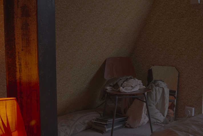 In the corner of a dark room, a chair is piled with clothing. Next to it on the ground is a mirror.
